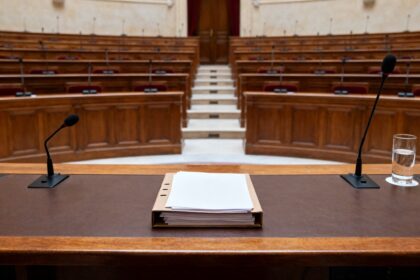 Salle de commission parlementaire vide, table d'audition avec deux micros, un dossier de documents et un verre d'eau.