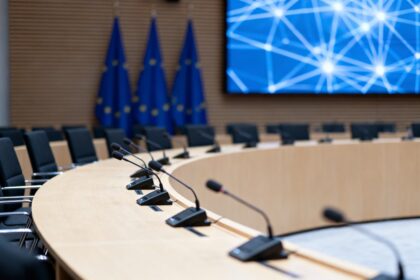 Salle de conférence institutionnelle européenne vide, micros alignés et écran affichant un réseau numérique