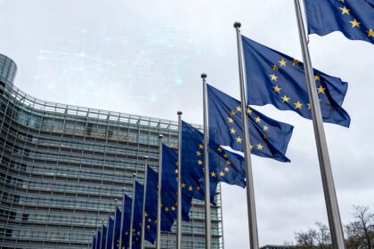 Rangée de drapeaux de l'Union européenne devant un bâtiment institutionnel vitré, sous un ciel gris