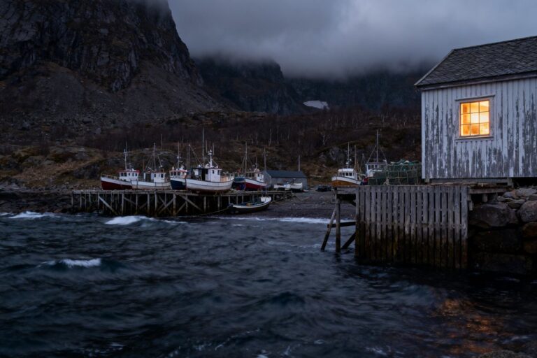Village de pêcheurs au crépuscule, bateaux amarrés le long d'un quai en bois et fenêtre éclairée face à une mer agitée