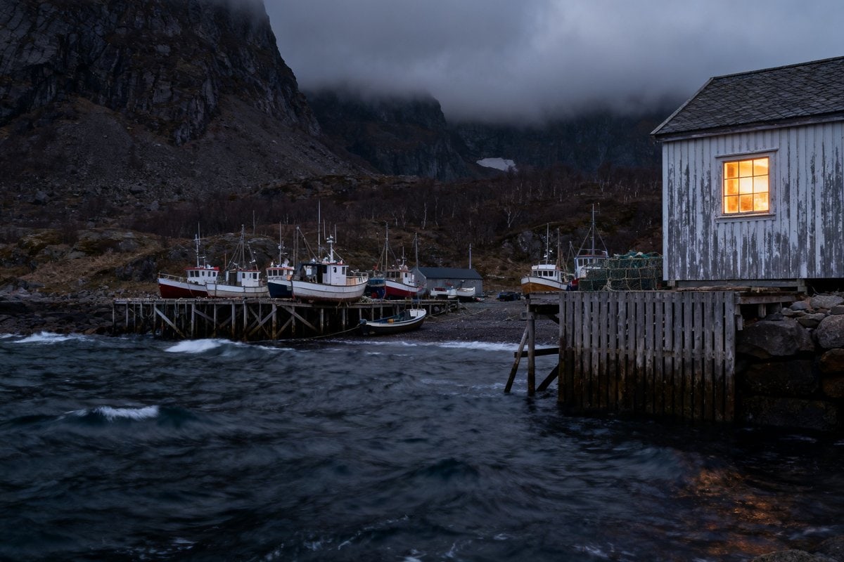 Village de pêcheurs au crépuscule, bateaux amarrés le long d'un quai en bois et fenêtre éclairée face à une mer agitée