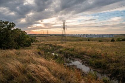 Prairie sèche traversée par un ruisseau et des pylônes, long bâtiment industriel bas à l'horizon