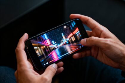 Deux mains tenant un smartphone qui diffuse une vidéo de rue nocturne aux néons sous la pluie