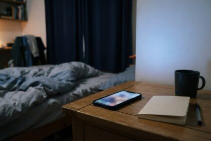 Smartphone allumé posé sur une table de chevet dans une chambre sombre, près d'un carnet fermé et d'un mug