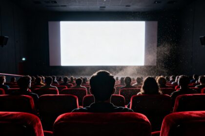 Salle de cinéma comble vue de dos, spectateurs en silhouette devant un écran blanc éclatant et des fauteuils rouges
