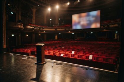 Salle de théâtre vide vue depuis la scène, sièges rouges, pupitre en bois et grand écran allumé.