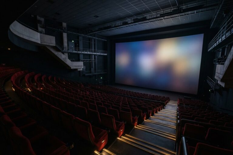 Grande salle de cinéma vide vue des rangs hauts, écran allumé face à des rangées de sièges rouges