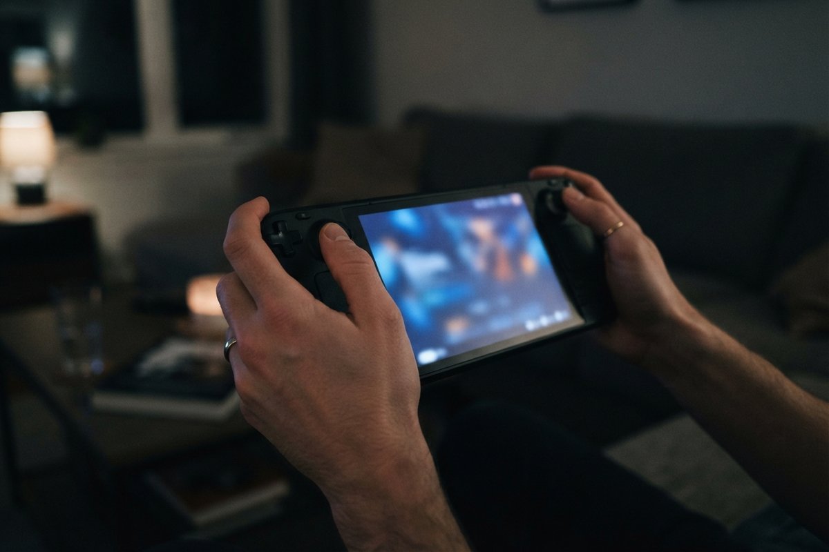 Deux mains tiennent une console de jeu portable dans un salon sombre, l'écran allumé éclaire les doigts