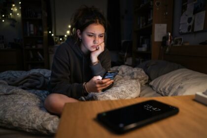 Adolescente assise sur son lit la nuit, les yeux sur son smartphone, un autre téléphone affichant 23:47 sur le bureau