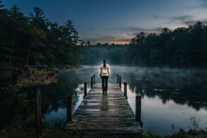 Femme de dos au bout d'un ponton sur un lac brumeux au crépuscule, près d'un panneau en bois Camp Crystal Lake