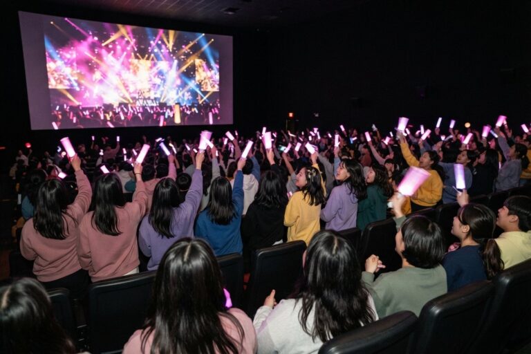Salle de cinéma comble, spectateurs brandissant des bâtons lumineux roses devant un écran aux lumières de concert