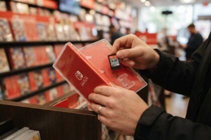 Mains tenant une boîte de jeu Switch rouge ouverte et une cartouche, devant des rayons de jeux dans un magasin